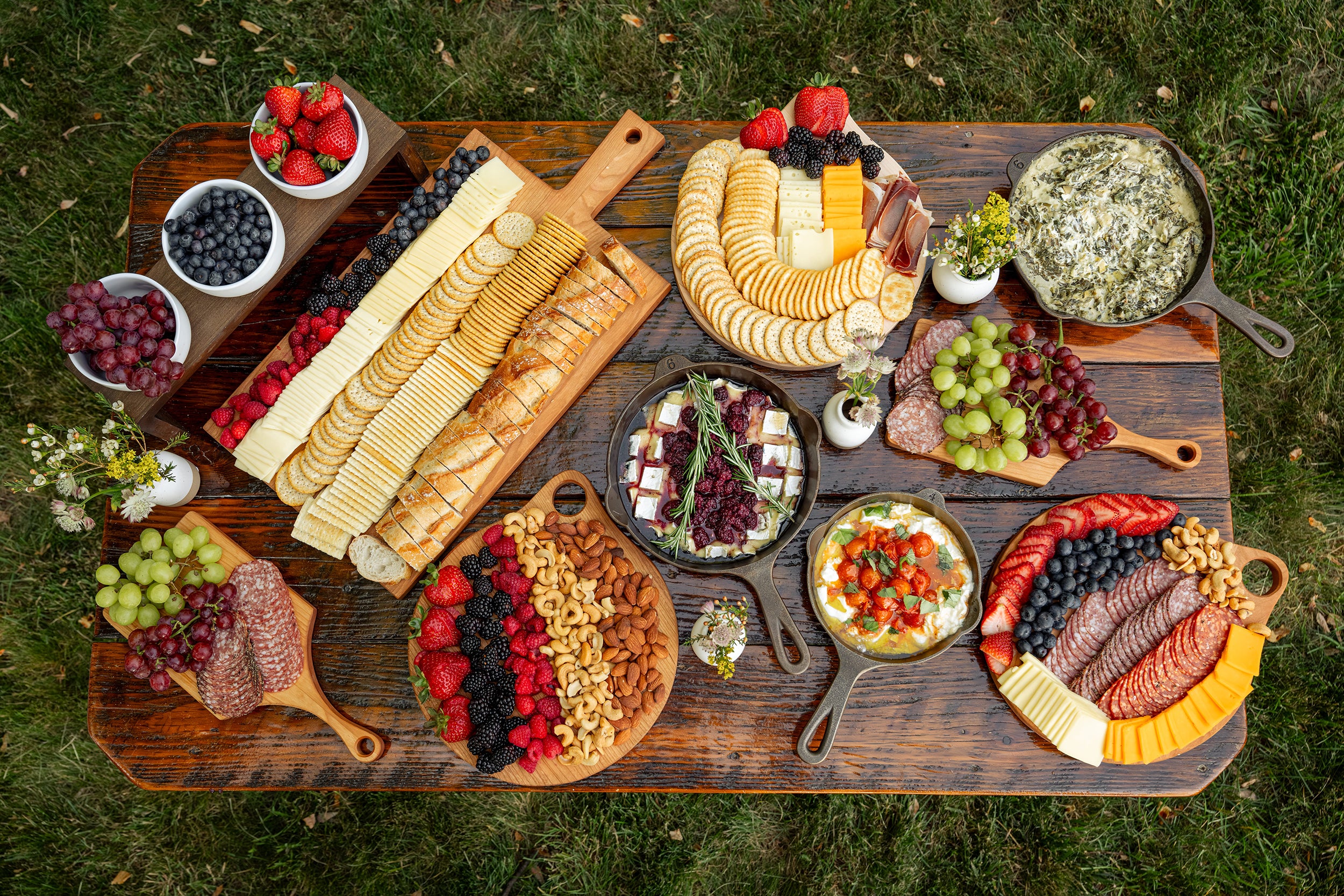 An overhead view of a large lancaster cast iron charcuterie and cheese board arranged on a wooden picnic table outdoors on grass, filled with sliced cheeses, crackers, meats, fruits, nuts, and dips.